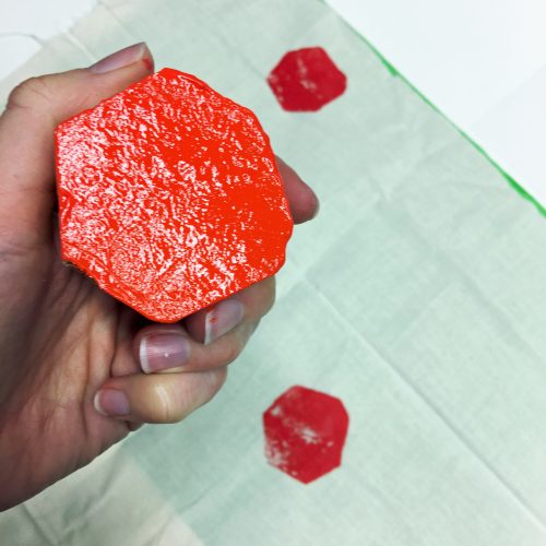 Potato half covered in red paint held by hand. Fabric with two red printed shapes sits in background
