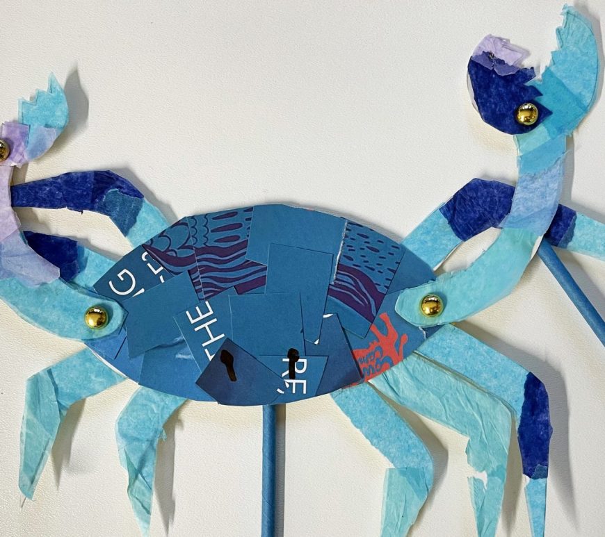 Blue crab puppet made of paper and collage materials on white background. It's claws and body have straws attached for moving the limbs in a shadow puppet style